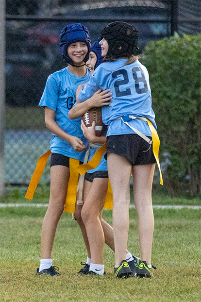 Three young girls in blue jerseys and protective helmets celebrate during a flag football game.